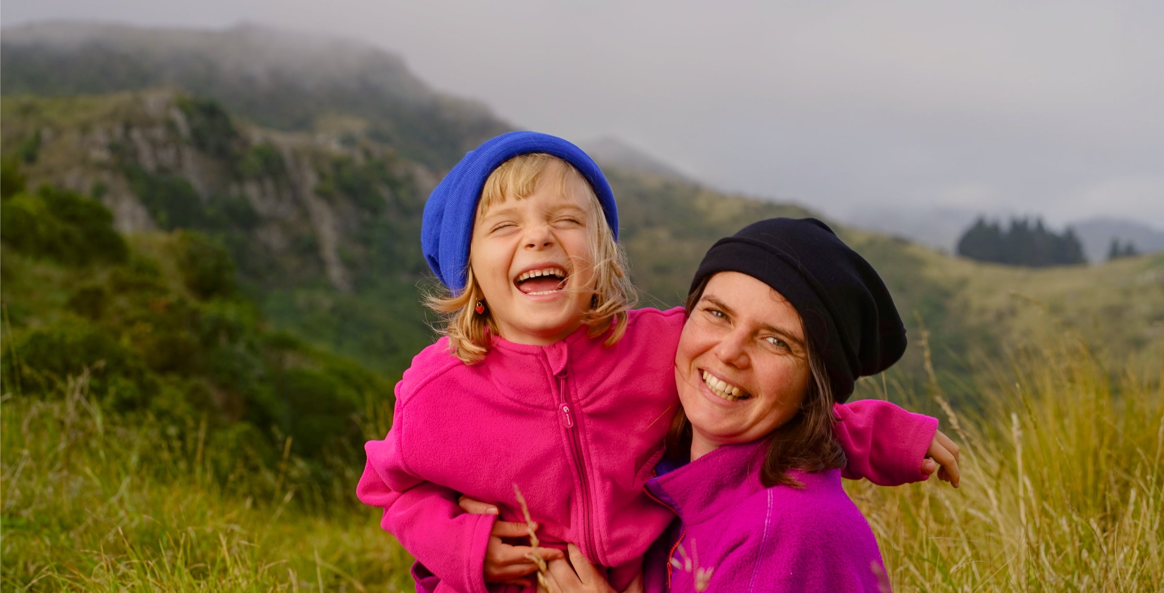 A woman and her daughter wearing lapis and black beanies in a hilly landscape while laughing.