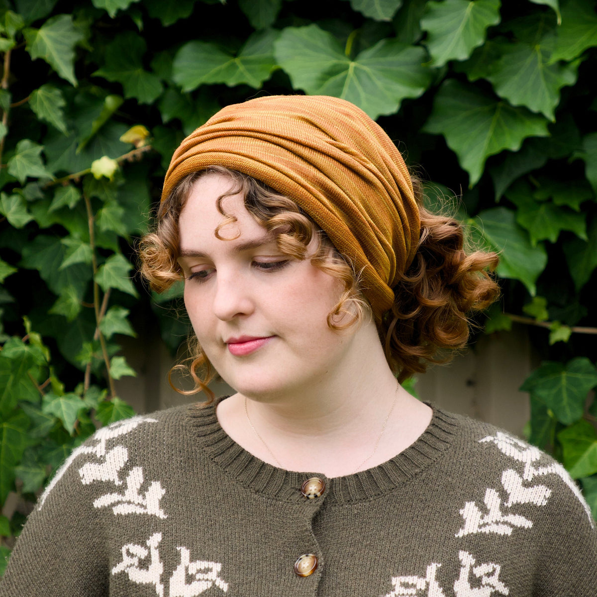 A woman wearing a mustard yellow fabric headwrap and a brown knit sweater with white leaf patterns, standing in front of a background of green ivy.