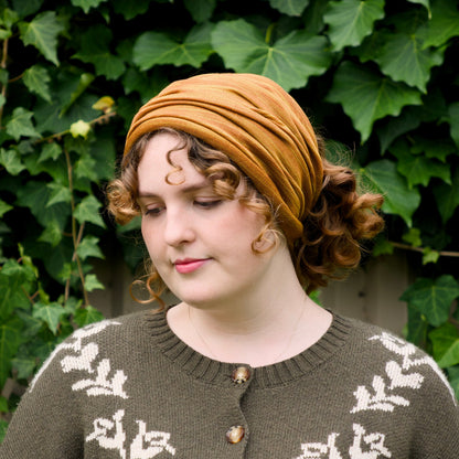 A woman wearing a mustard yellow fabric headwrap and a brown knit sweater with white leaf patterns, standing in front of a background of green ivy.