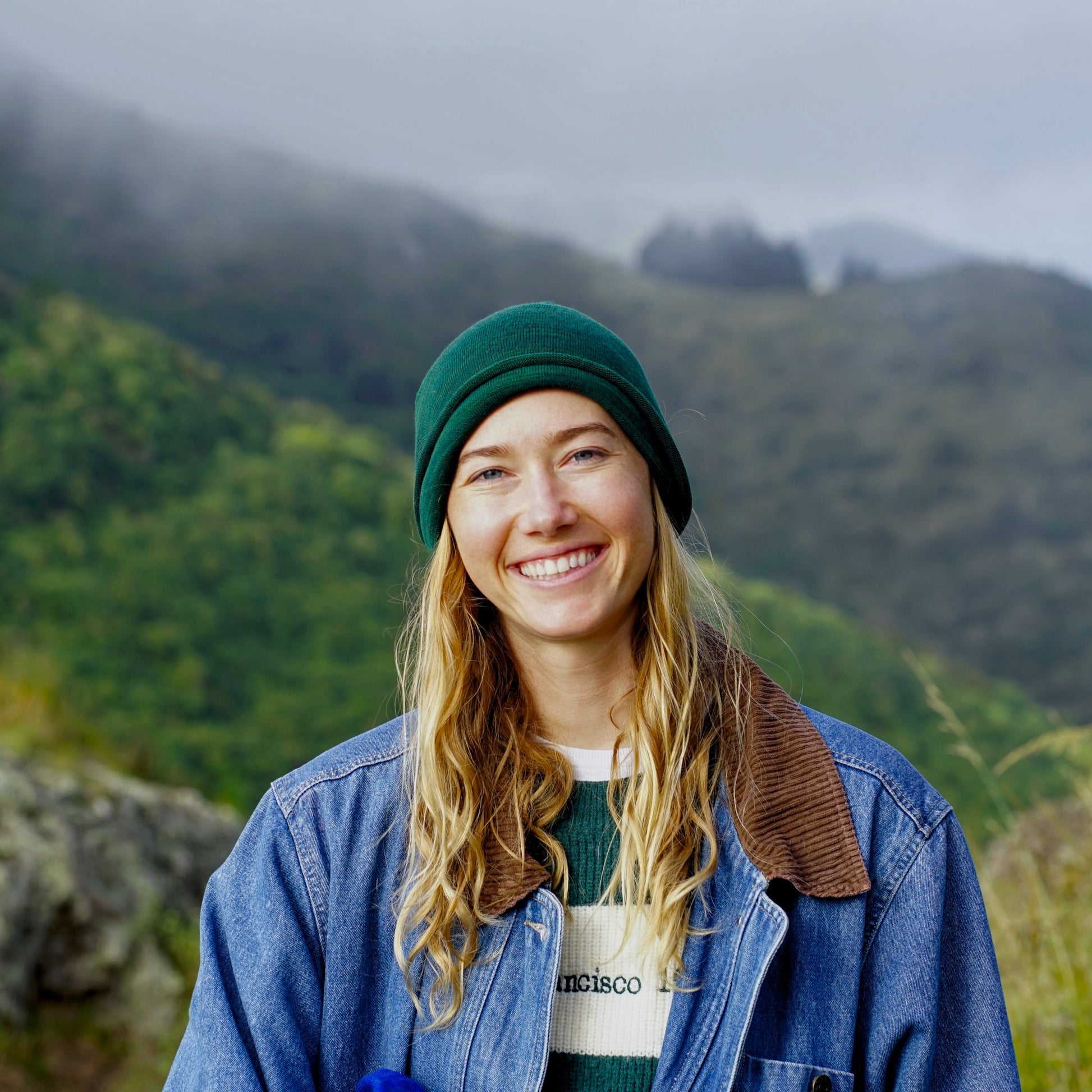 Person wearing a green beanie and blue jacket with a mountainous background